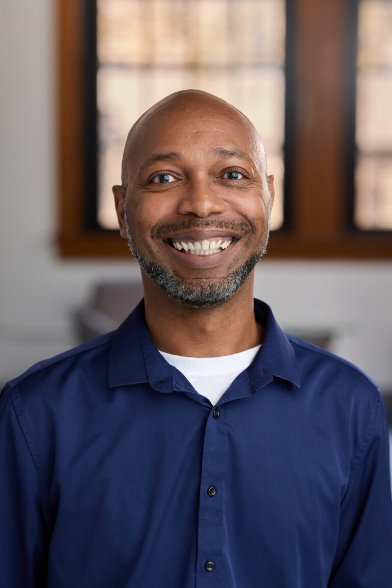 Close-up of a smiling man with a bald head, blue eyes, and a short beard wearing a navy shirt.