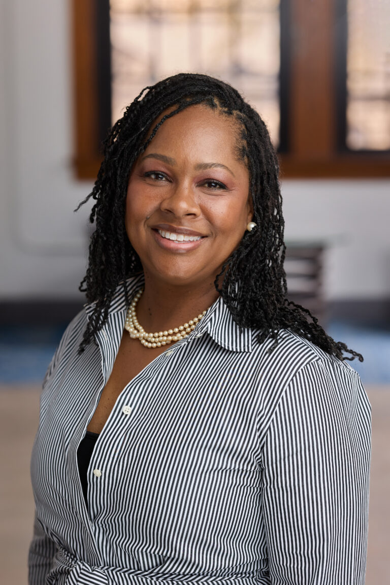 Portrait of a smiling woman wearing a black-and-white striped blouse and pearl necklace indoors.