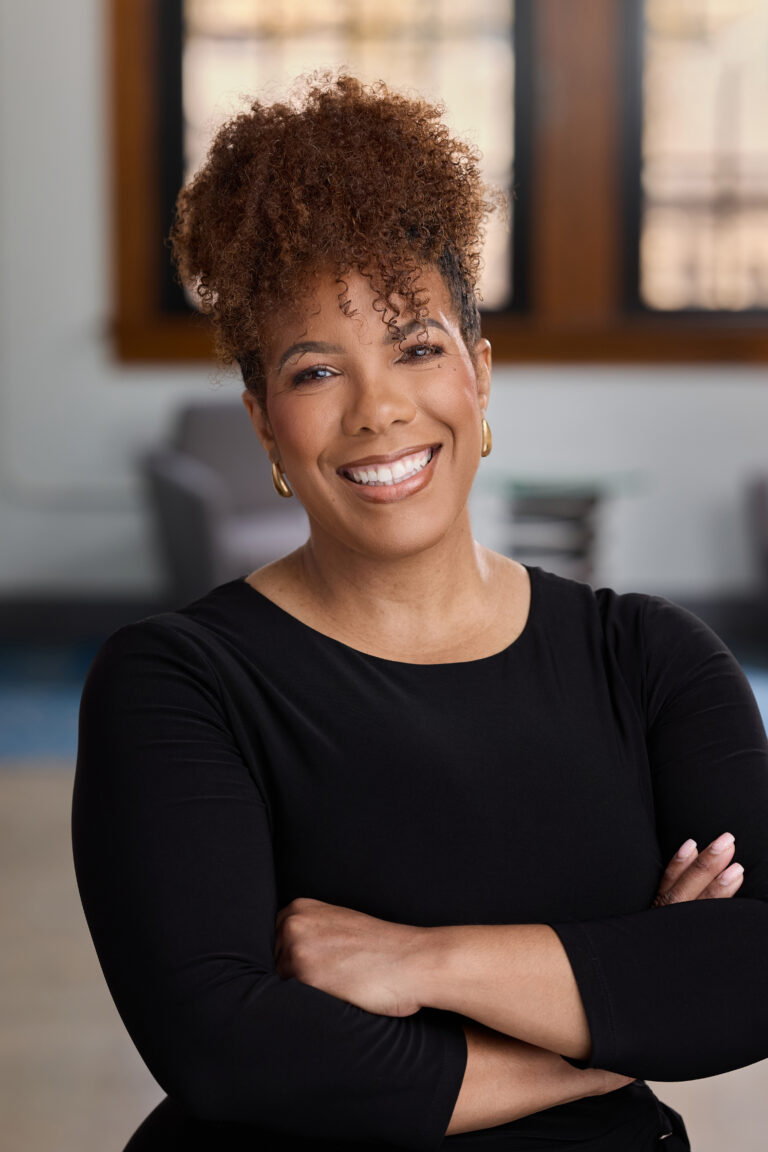 Smiling woman with curly hair wearing a black top, arms crossed, indoors in soft light.