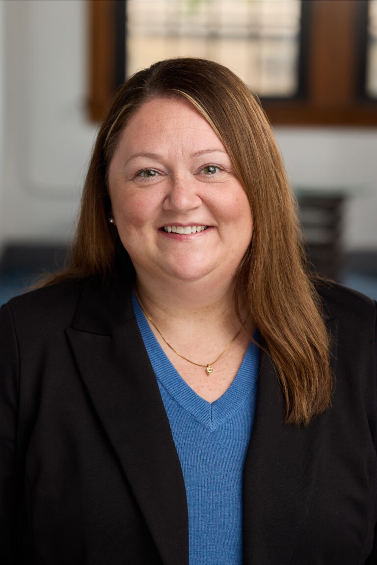 Close-up portrait of a smiling woman with brown hair, wearing a blue sweater and black blazer indoors in an office setting.