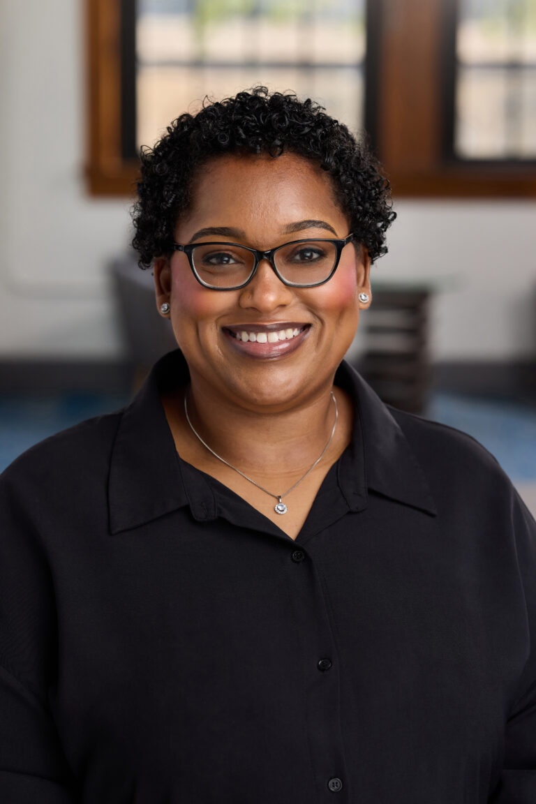 Smiling woman with short curly hair and glasses, wearing a black button-up shirt and necklace, indoor background blur