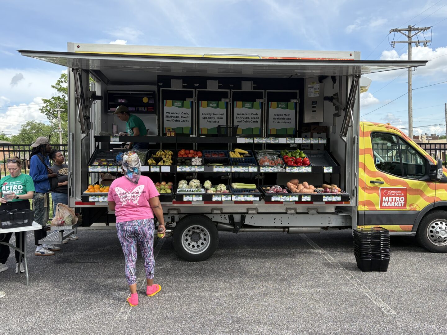 Open-air mobile grocery truck offering fruits and vegetables, with staff assisting customers in a parking lot.
