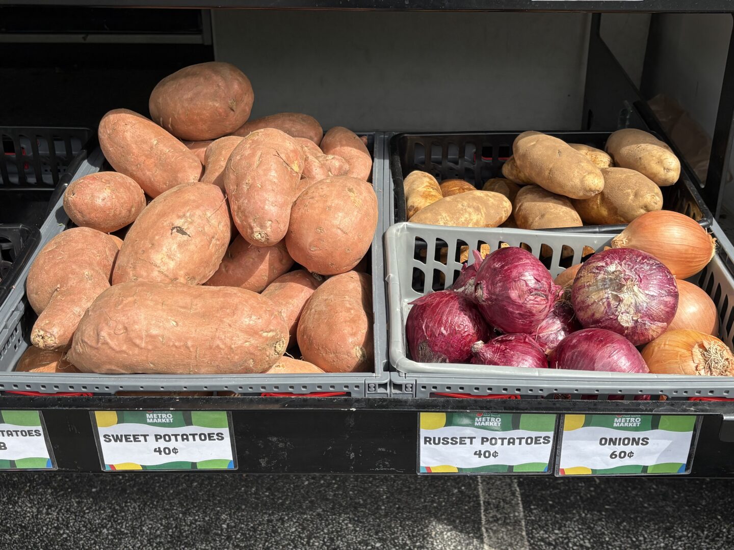 Pile of orange sweet potatoes in a gray crate at a market, with neighboring russet potatoes and red onions in crates nearby.