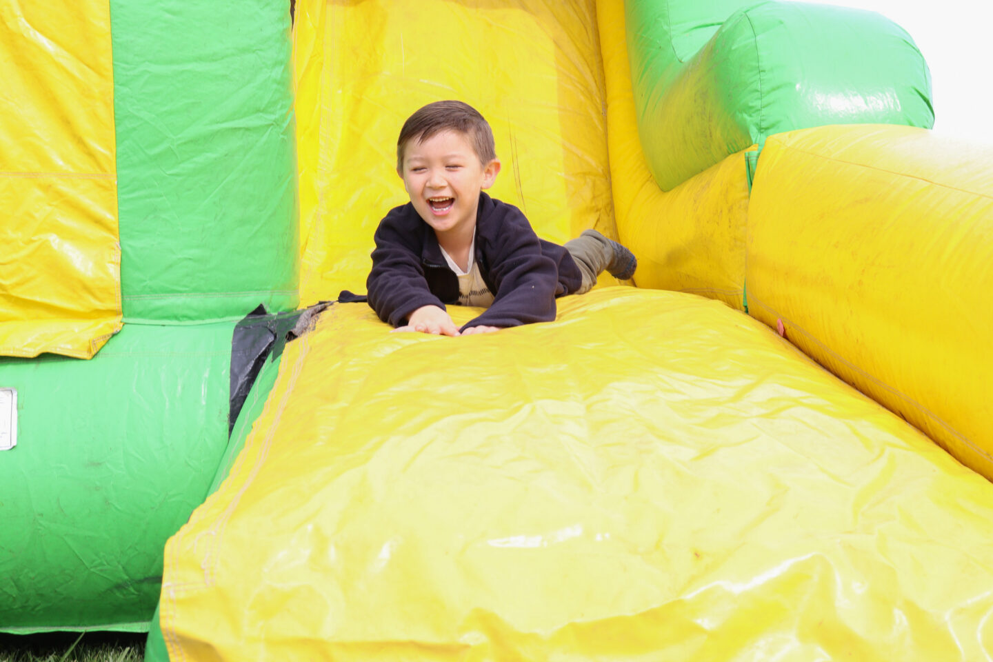 Boy in a dark jacket laughing as he slides down a bright yellow and green inflatable structure.