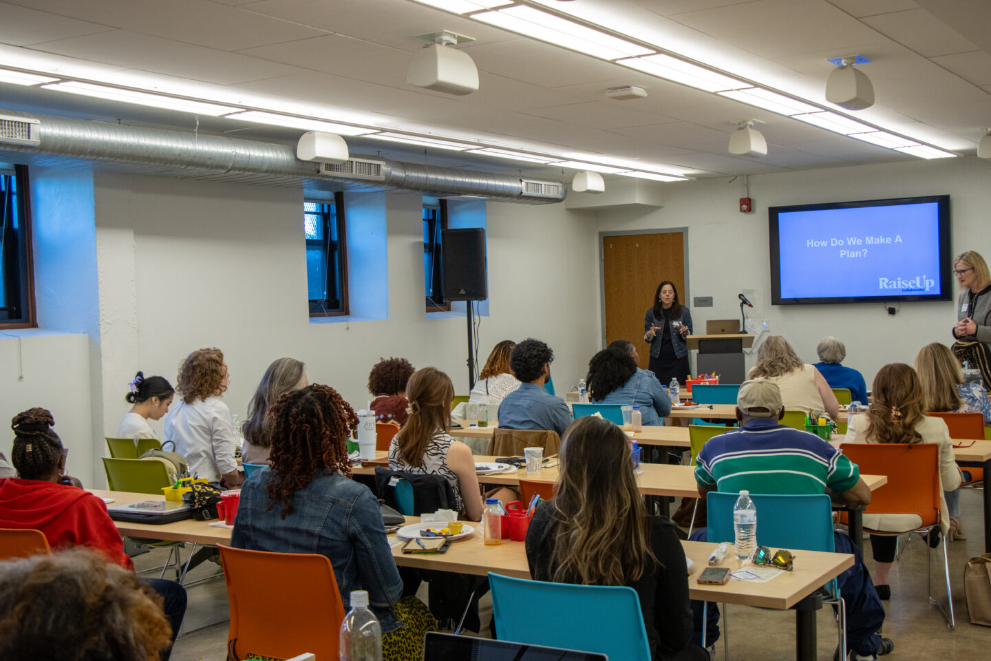 Presenter at a podium speaking to an audience in a classroom; screen reads 'How Do We Make A Plan?'.