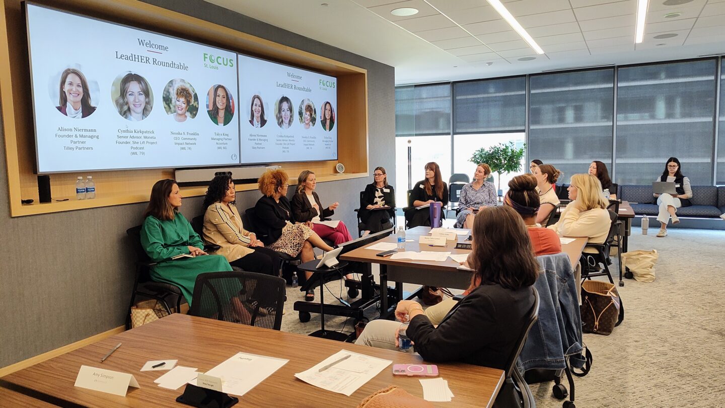 Panel discussion in a bright conference room; large screens show LeadHER Roundtable and Focus St. Louis logos with speaker headshots on display.