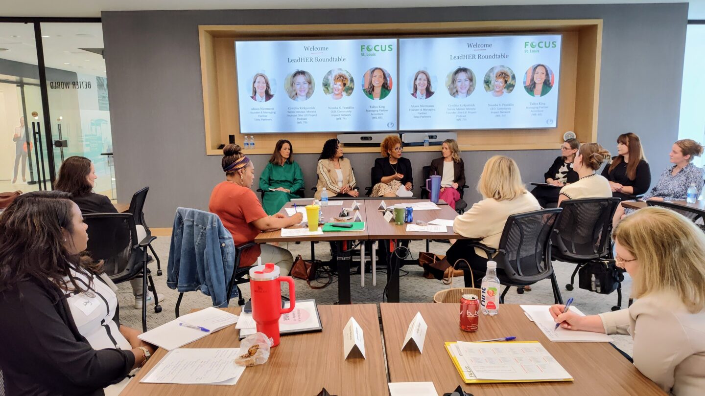 Group of professionals seated around a conference table during a roundtable discussion, with presentation screens on the wall behind them.