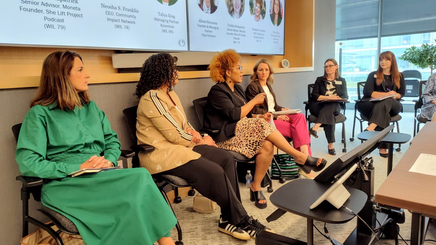 Six women seated in a row on a panel in a conference room, some with notebooks, with bio slides visible on screens behind.