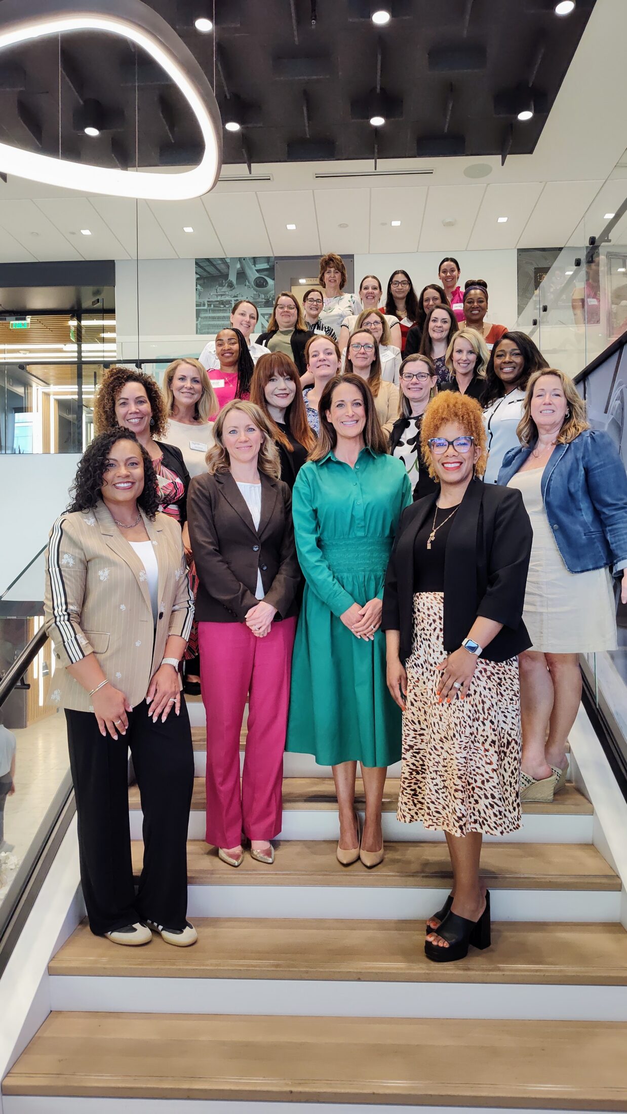 Group of diverse women in business attire posing on a staircase in a modern office lobby.