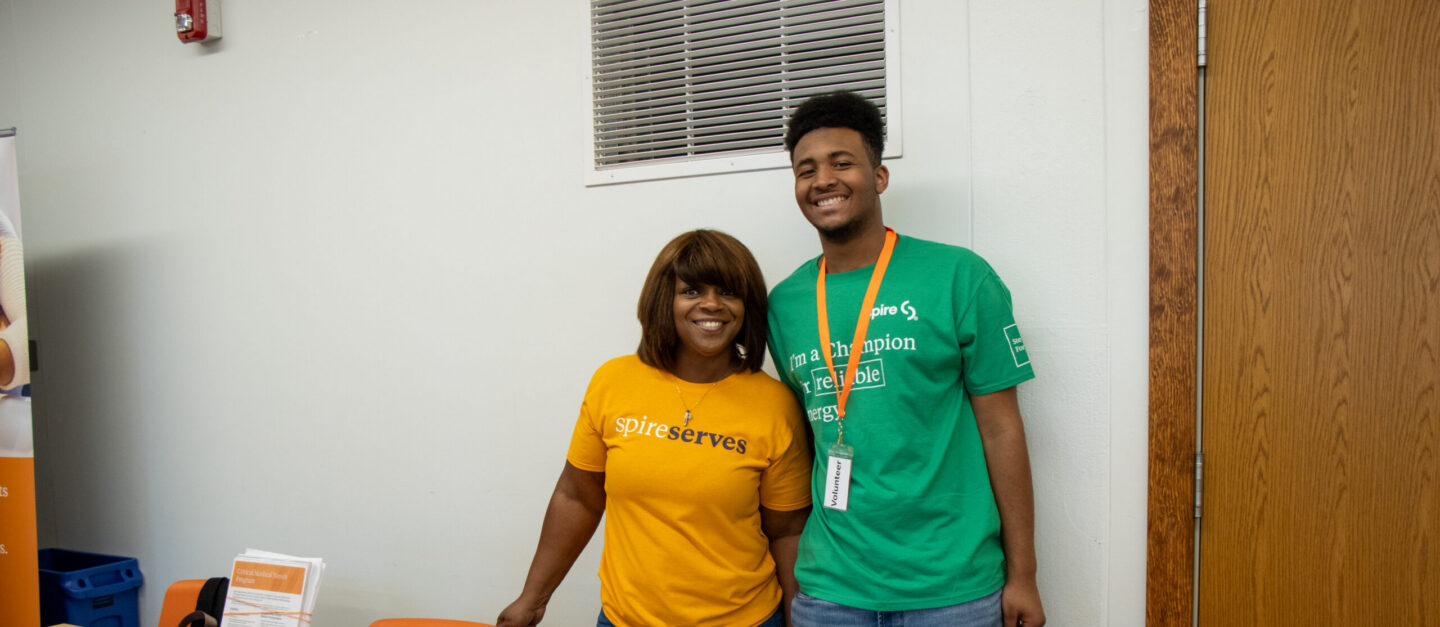 Two volunteers stand behind an orange table at a community event, smiling at the camera (one in a yellow shirt that says 'spire serves' and the other in green with an orange lanyard).