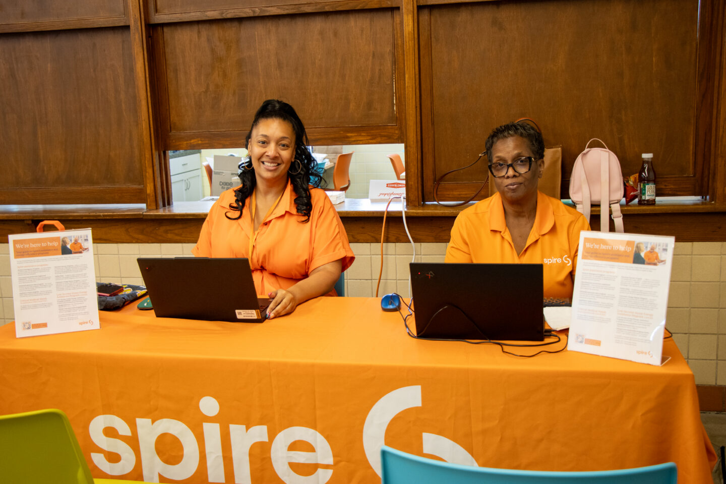 052826_SpireEvent-20 - Community Impact Network Two smiling women in orange uniforms sit at a desk with laptops in a help desk or reception area.