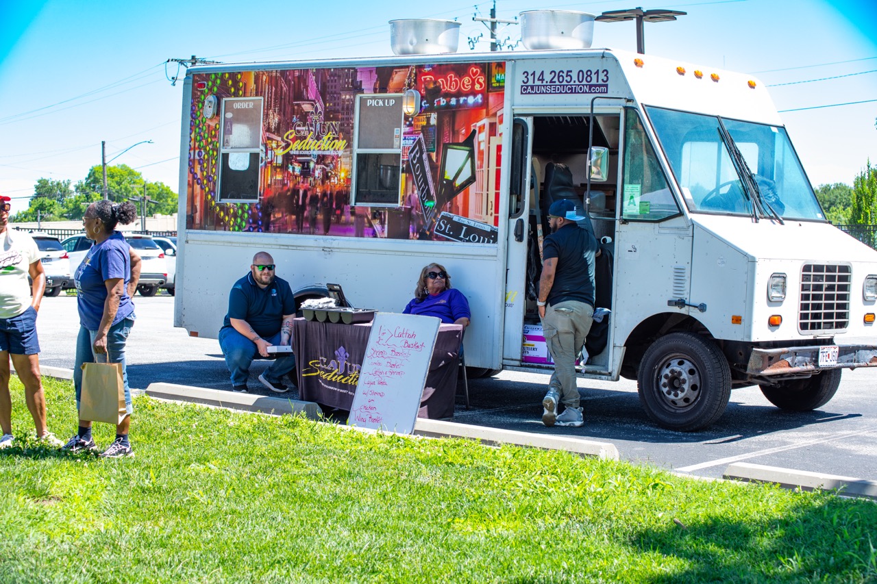1W3A6787 Large - Community Impact Network White Cajun food truck with bright city artwork, logo reading Cajun Seduction, open serving window, a man entering the truck, and two people seated at a table outside.