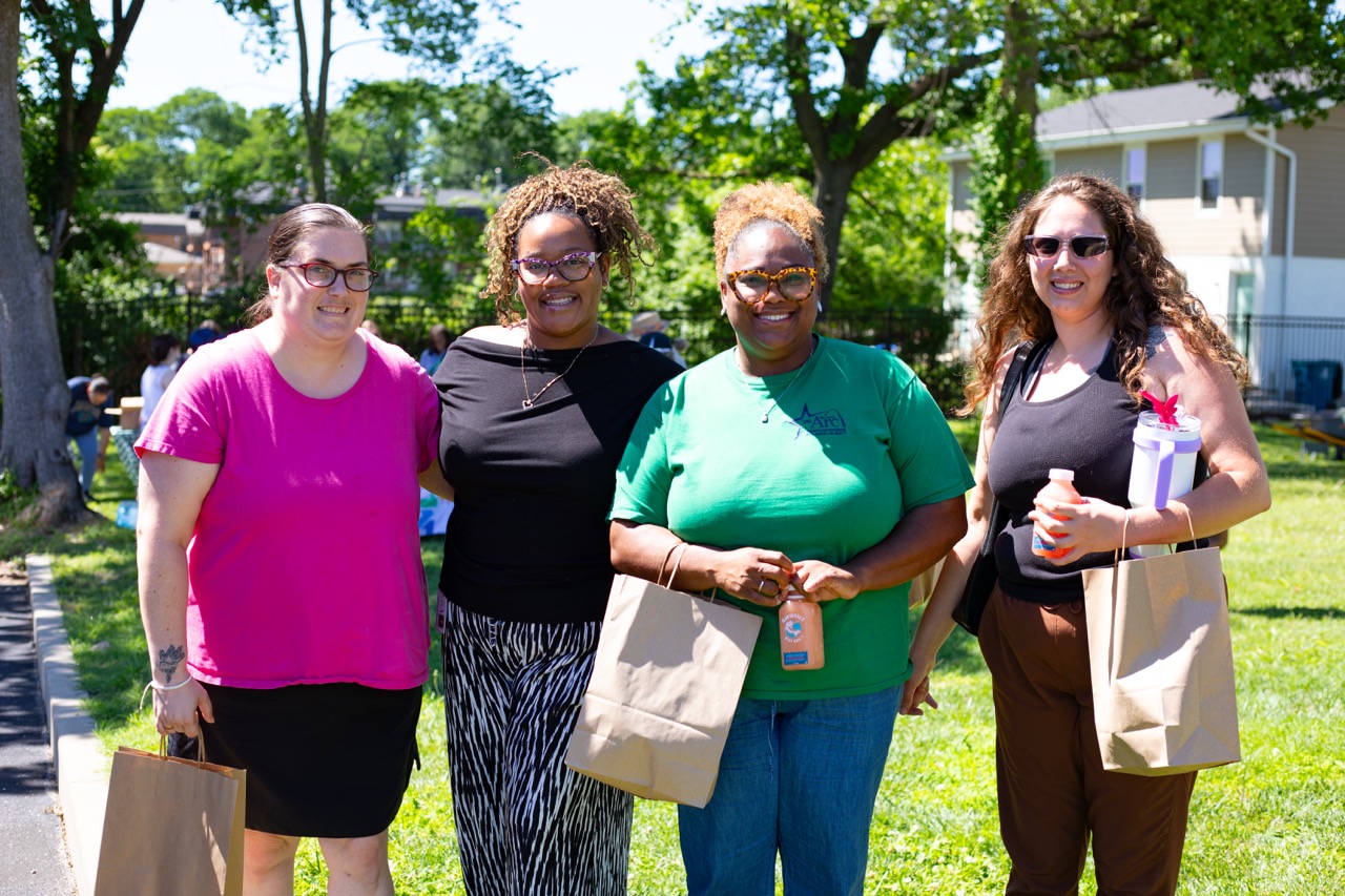 1W3A6836 Large - Community Impact Network Four women posing together outdoors on a sunny day, each holding a brown paper bag.