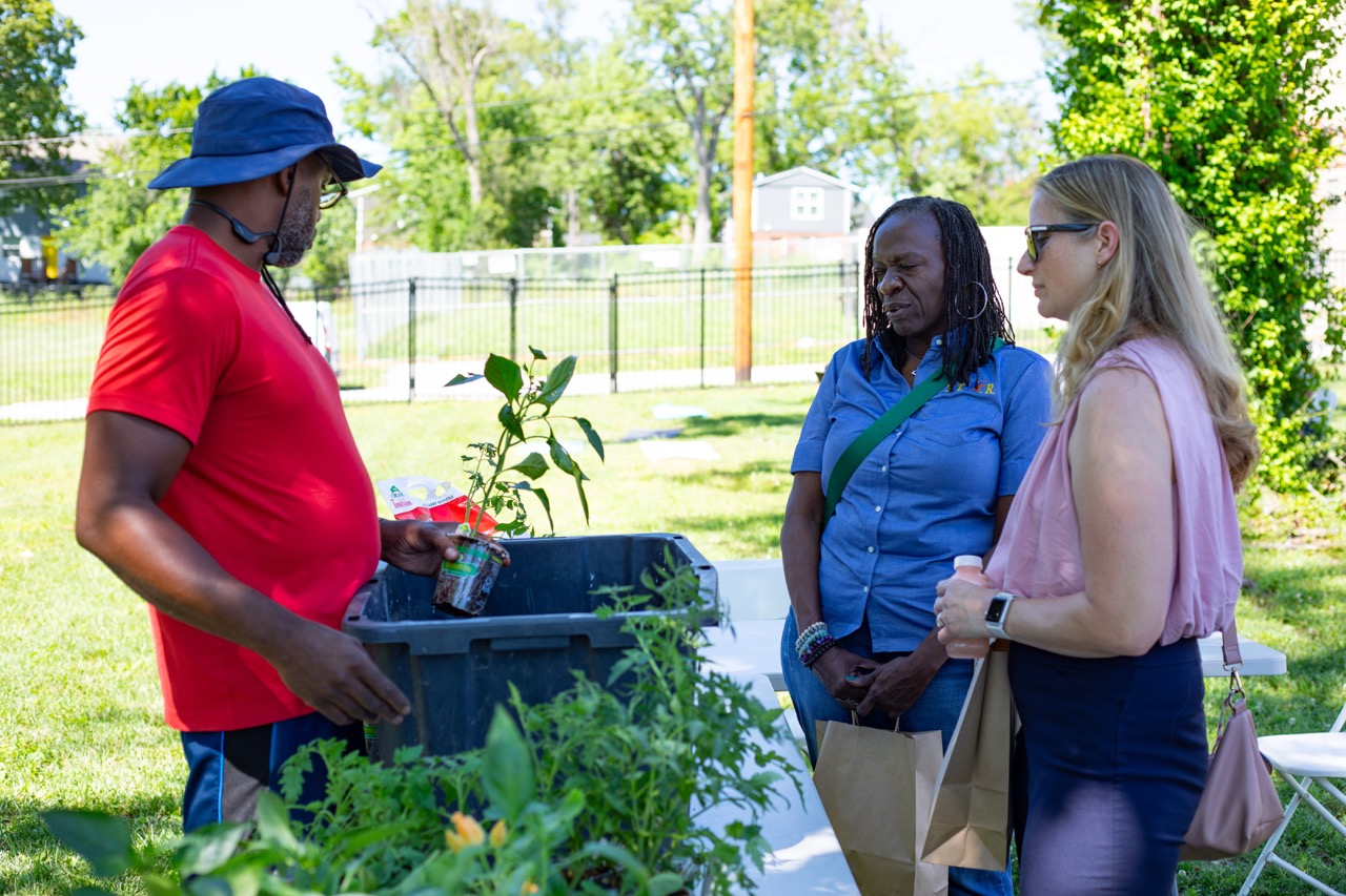 1W3A6852 Large - Community Impact Network Man in a red shirt and blue hat shows a plant to two women at a community garden table outdoors.