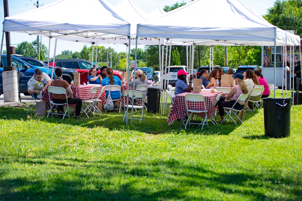 1W3A6951 Large - Community Impact Network Community picnic under white tents with groups of people at red-and-white checkered tables outdoors on a sunny day.