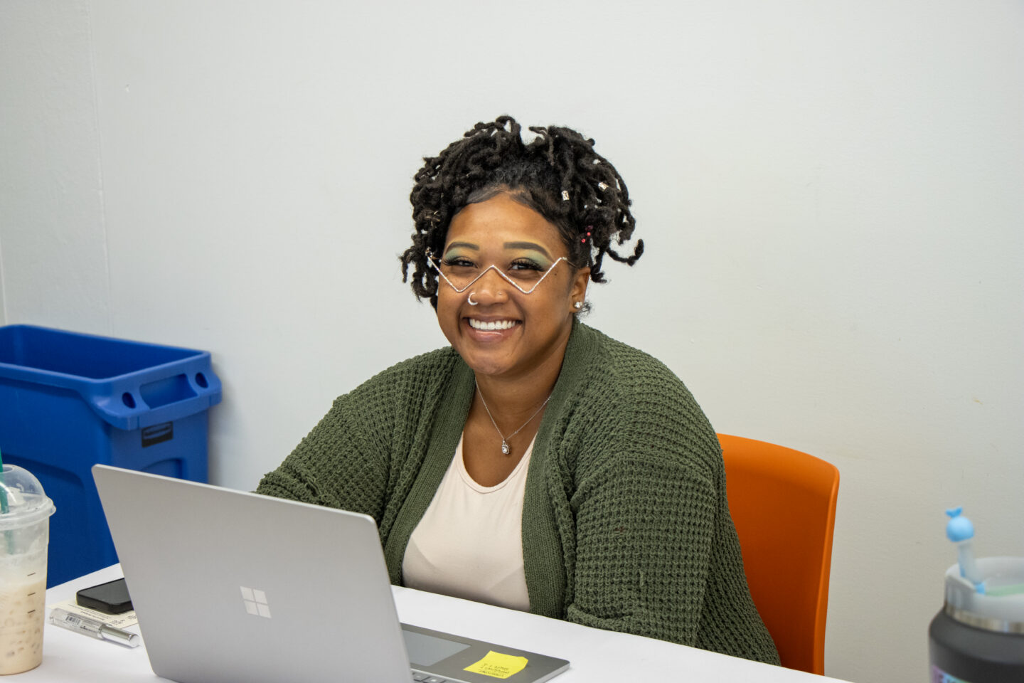 Smiling woman with decorative white eyewear face paint uses a laptop at a desk in a bright office, with a blue bin and orange chair nearby.