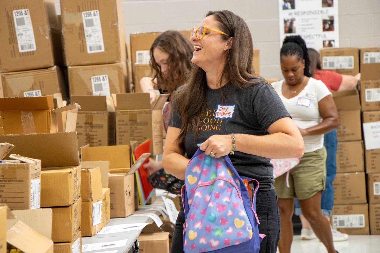 Woman with orange glasses laughing among stacked cardboard boxes, holding a heart-pattern backpack.