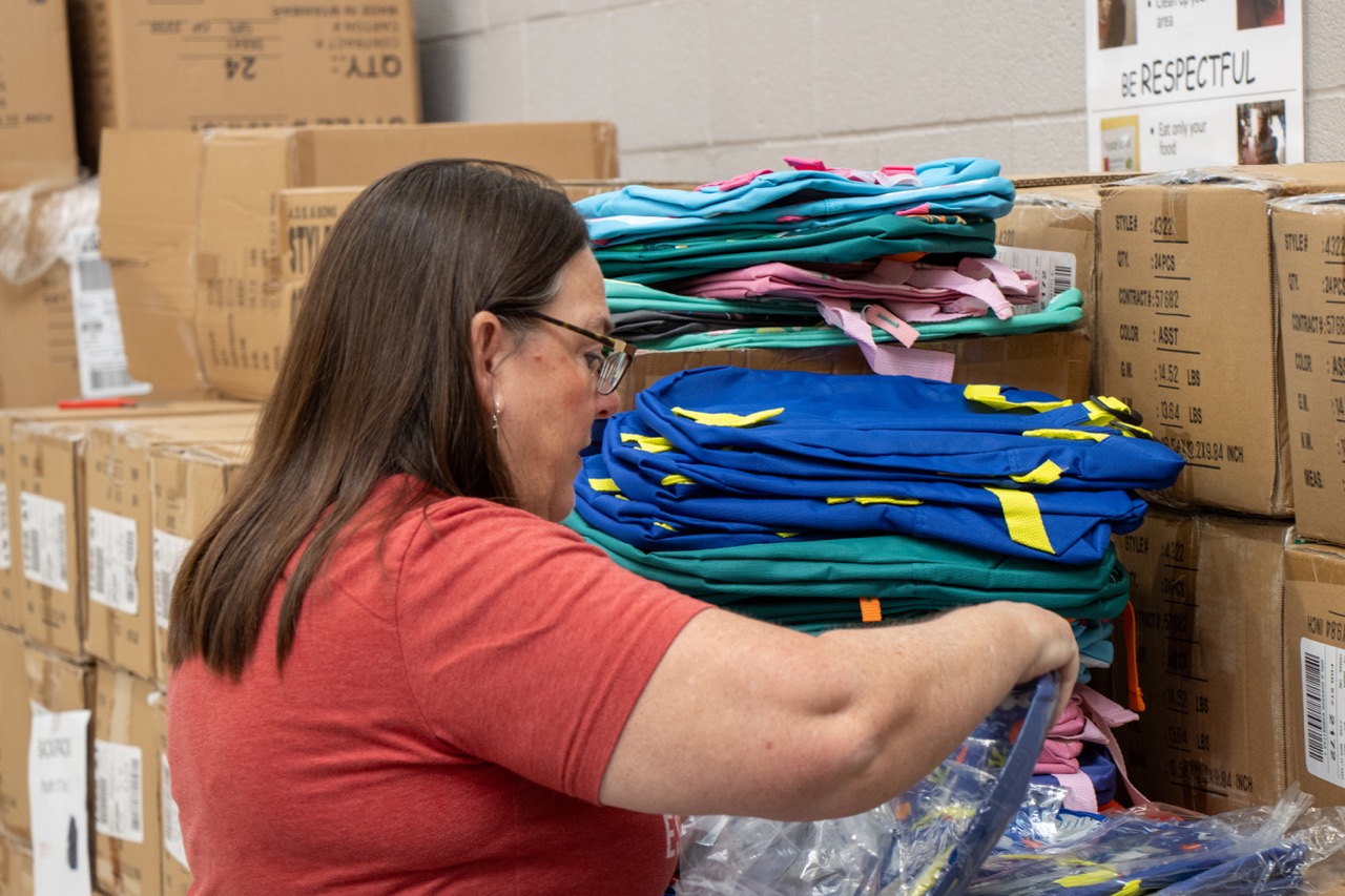 Woman in a red shirt and glasses sorts colorful folded clothing in a storage room with cardboard boxes.