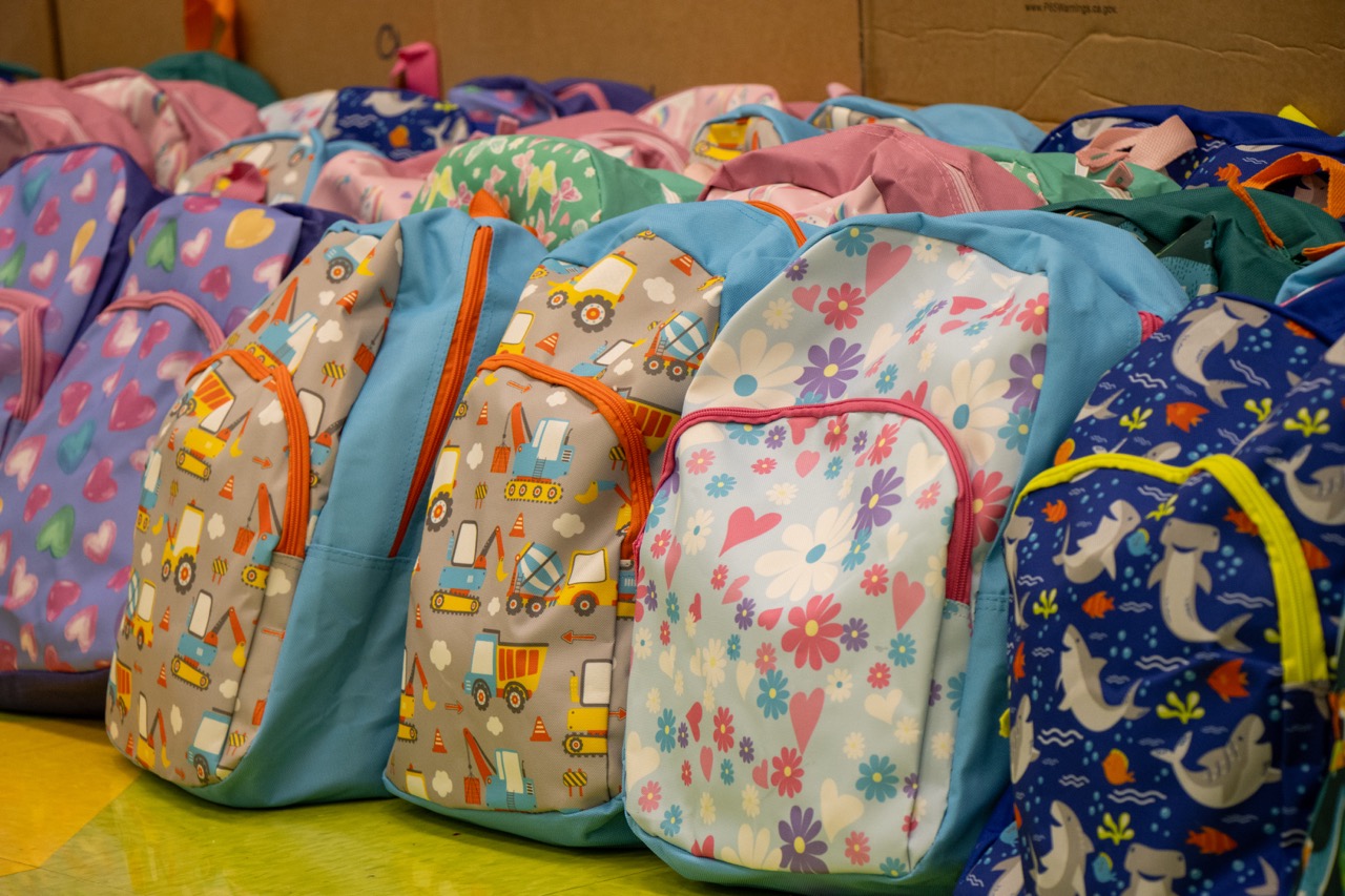 Row of colorful children's backpacks with patterns including hearts, flowers, construction trucks, and sea creatures.