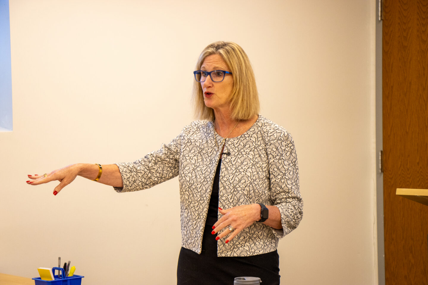Woman with blue glasses speaking and gesturing with her arms in a classroom, wearing a patterned blazer and black dress.
