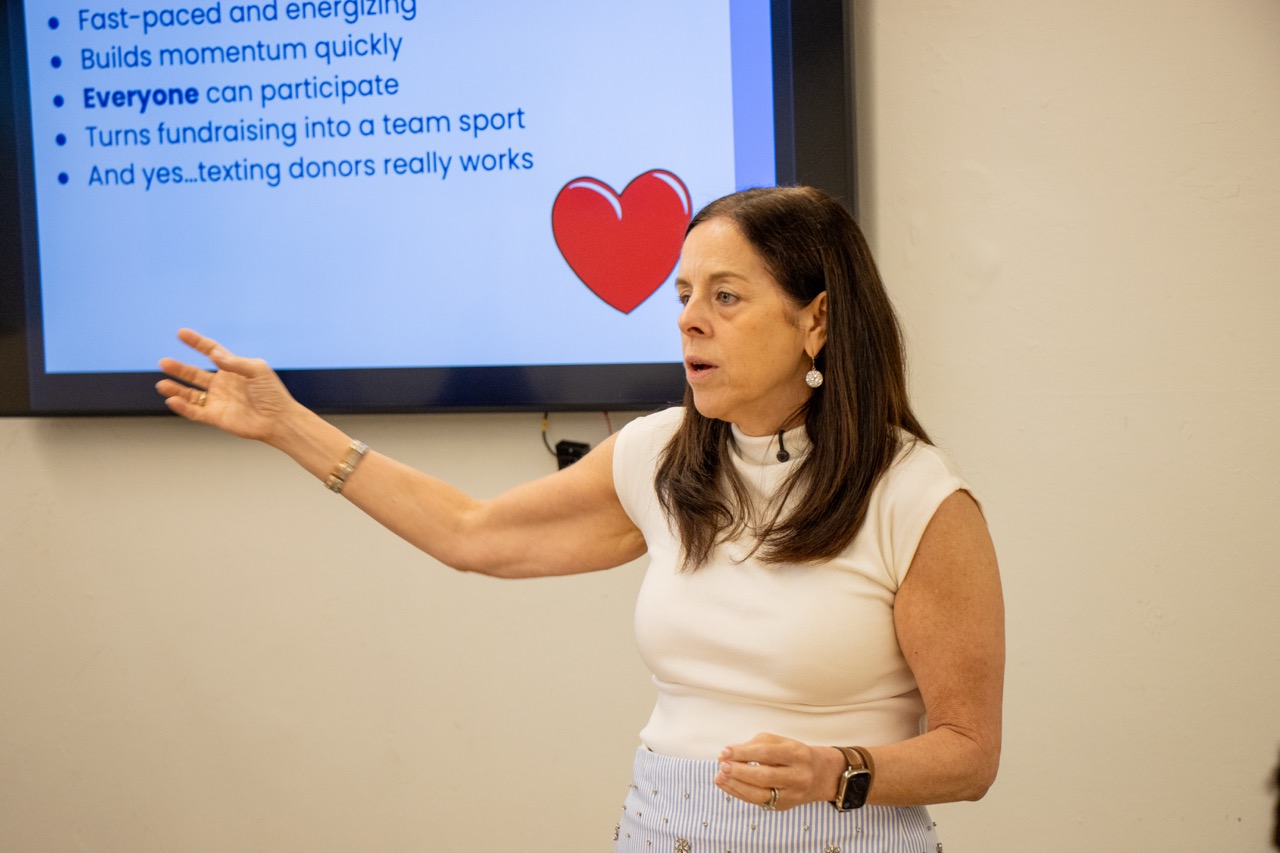 Presenter in a white sleeveless top gestures toward a slide with a large red heart and bulleted points.