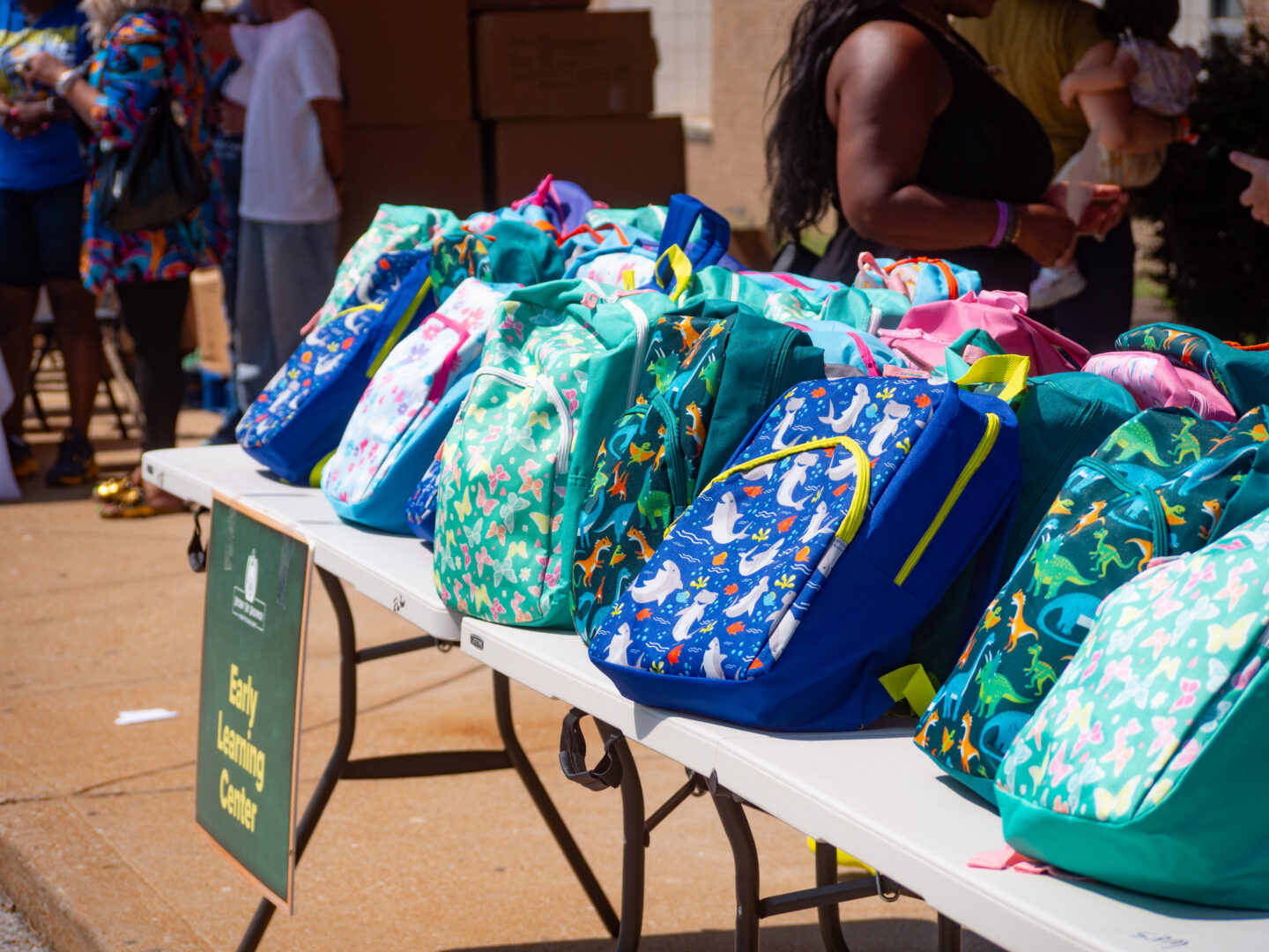 P4443581 - Community Impact Network Rows of colorful backpacks with printed designs on a table at an outdoor event, with people standing in the background.