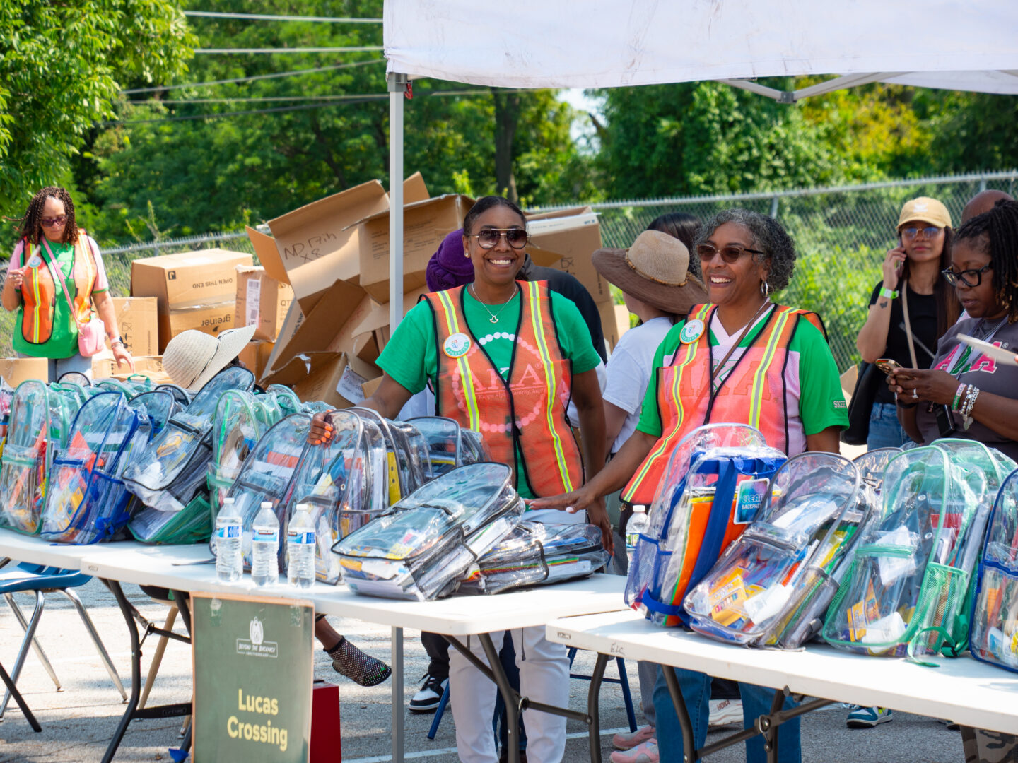 P4443633 - Community Impact Network Volunteers in neon vests handing out cleared backpacks and supplies at an outdoor charity event with boxes in the background, smiling volunteers.