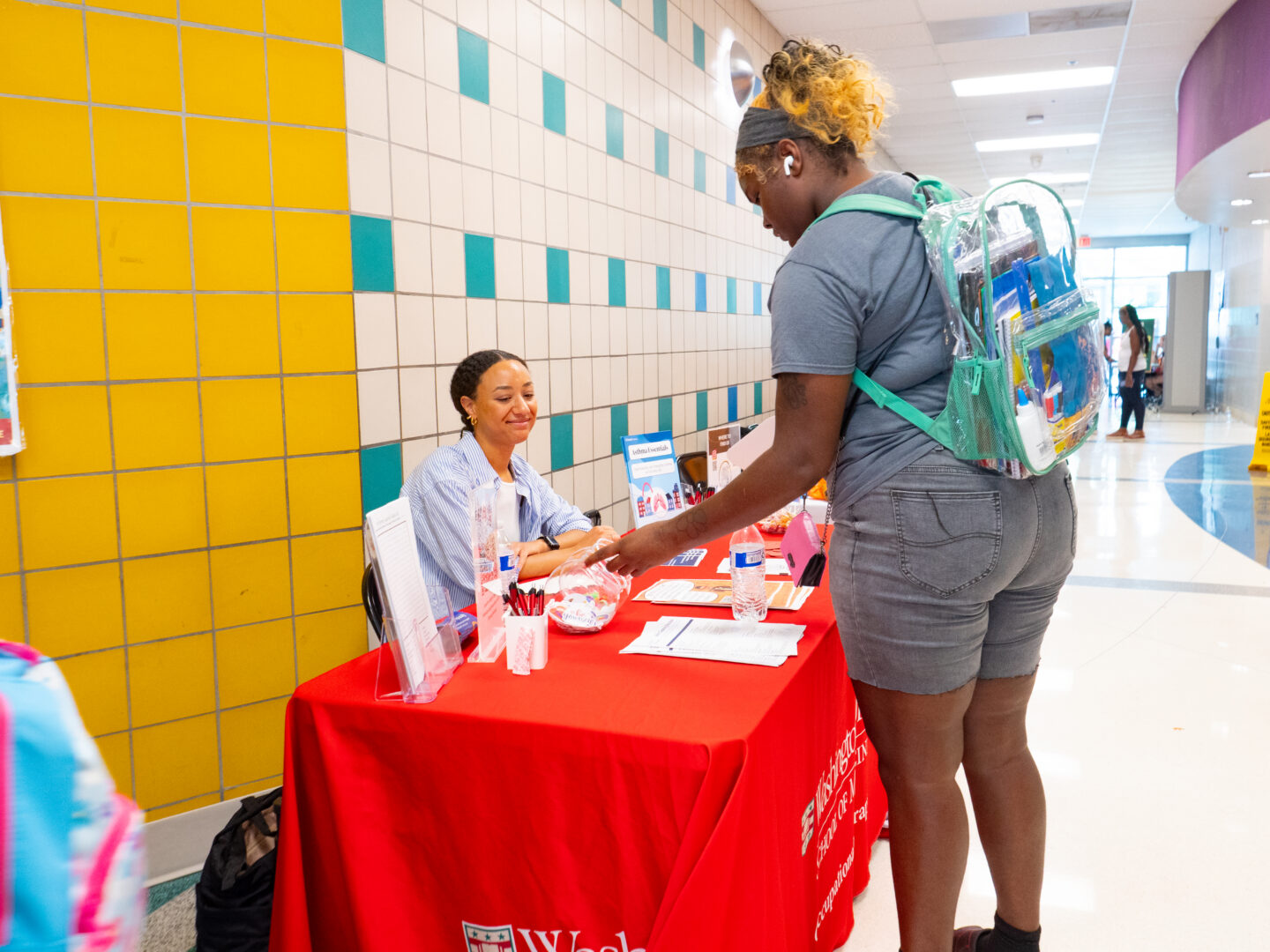 P4443671 - Community Impact Network Staff member at a red information desk assisting a customer with a clear backpack; yellow and teal tiled wall behind them.
