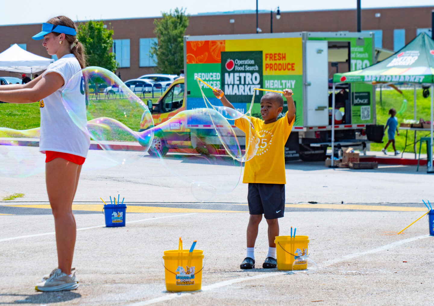 Girl wearing a blue visor and white shirt blows large bubbles as a boy in a yellow shirt watches nearby, yellow buckets on the ground at an outdoor event.