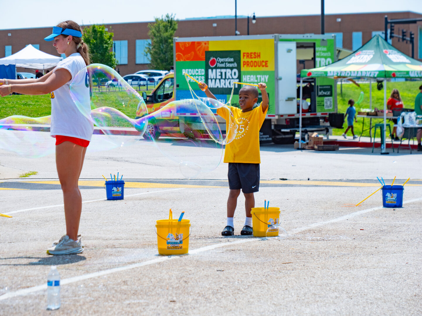 Girl wearing a blue visor blows giant bubbles while a boy in a yellow shirt creates bubbles outdoors at a festival.
