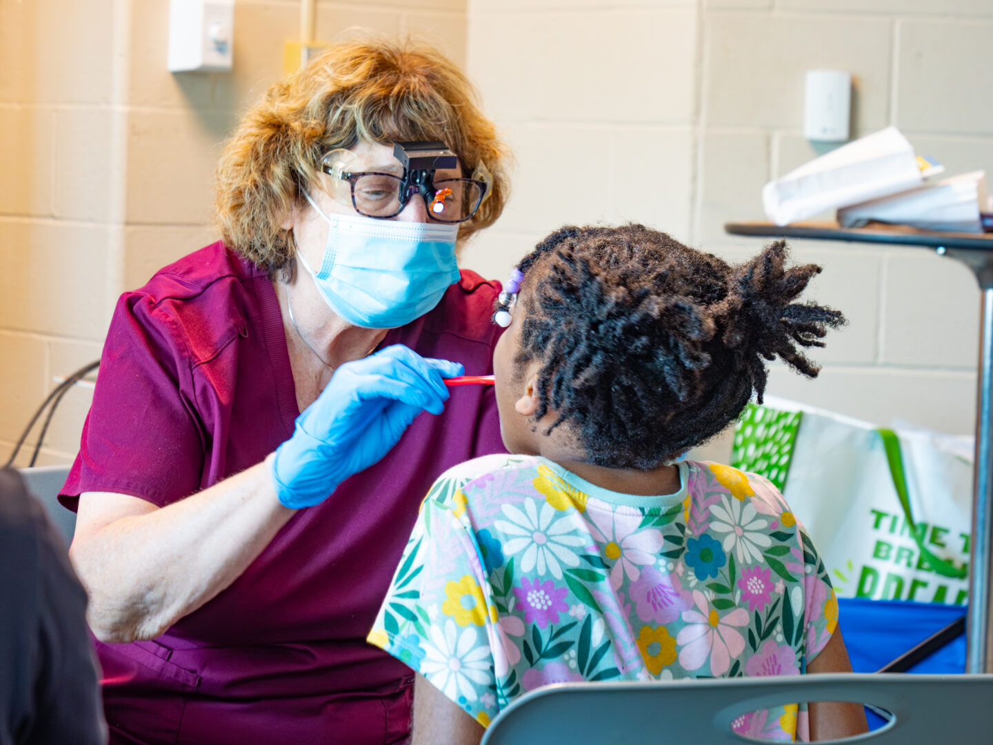 P4443919 - Community Impact Network Clinician in a mask and magnifying headlamp, wearing blue gloves, examines a child with a red dental tool in a clinic setting.