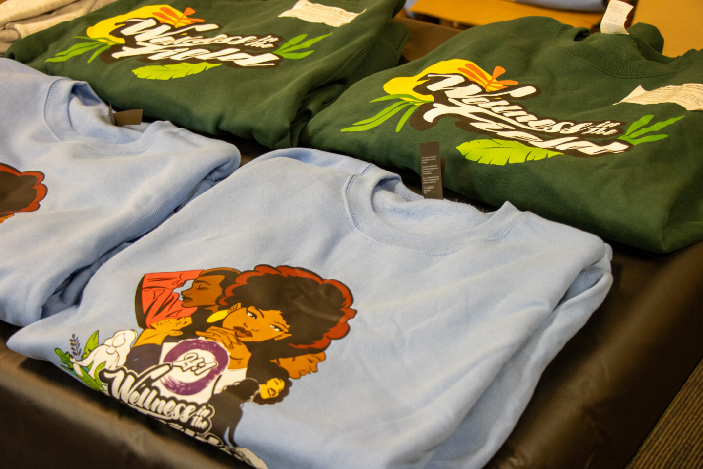 Pile of folded graphic T-shirts in blue and green, featuring colorful retro illustrations of people and bold white script text.