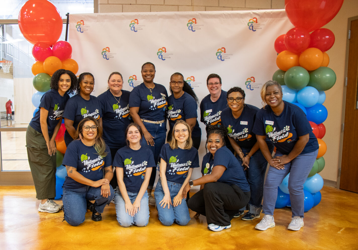 Group of diverse volunteers in navy T-shirts posing together at an indoor wellness event, with colorful balloon pillars on either side.