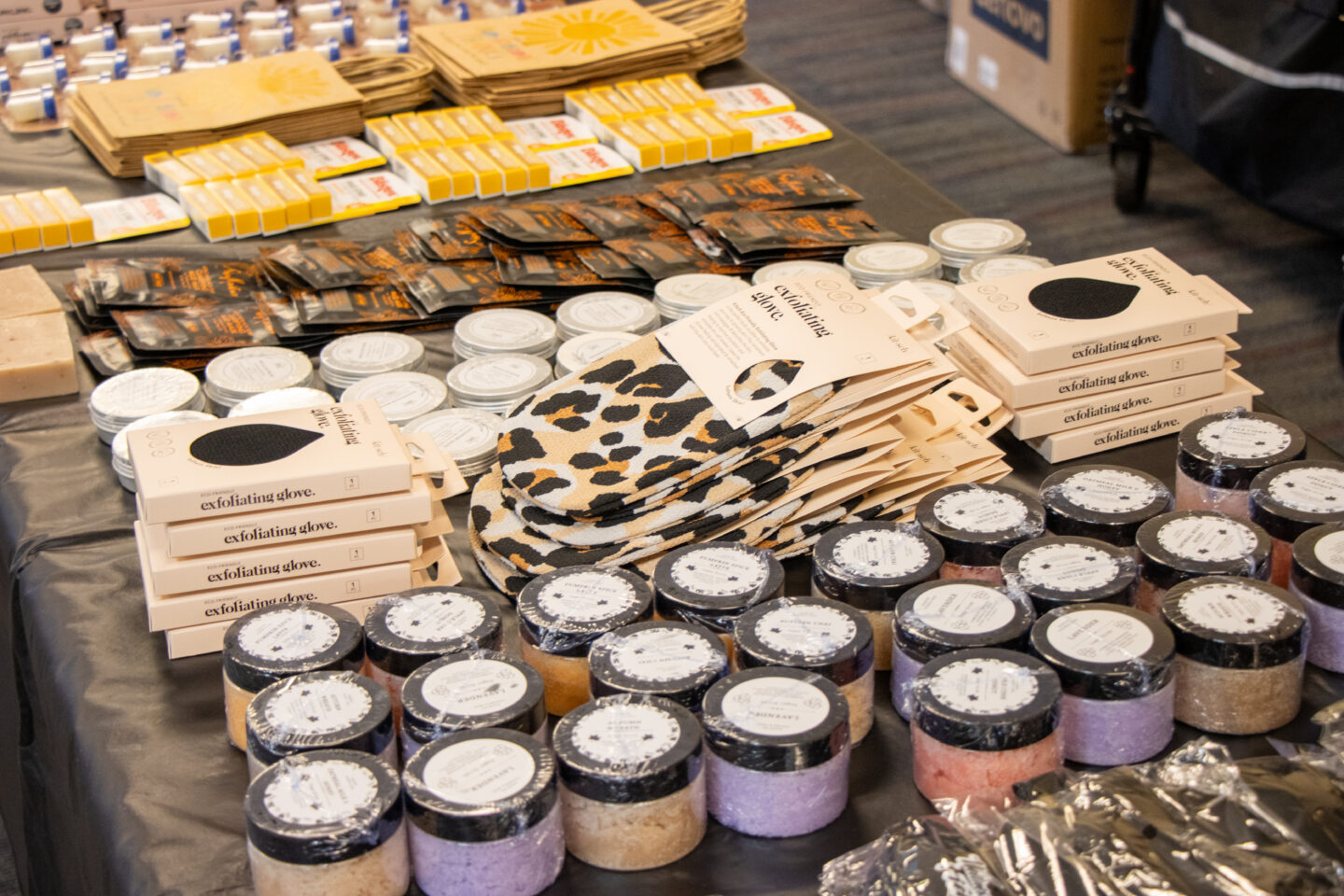 Display table of personal care items including exfoliating gloves boxes and assorted bath products like scrubs in jars and wrapped soaps on a black tablecloth