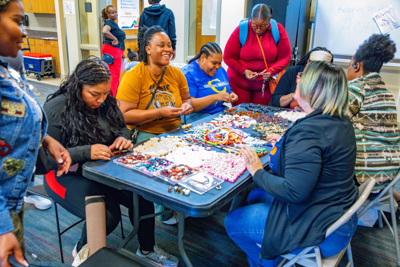 1W3A6233 Large - Community Impact Network Group of people seated around a table making and sorting colorful bead bracelets together at a community event in a classroom.