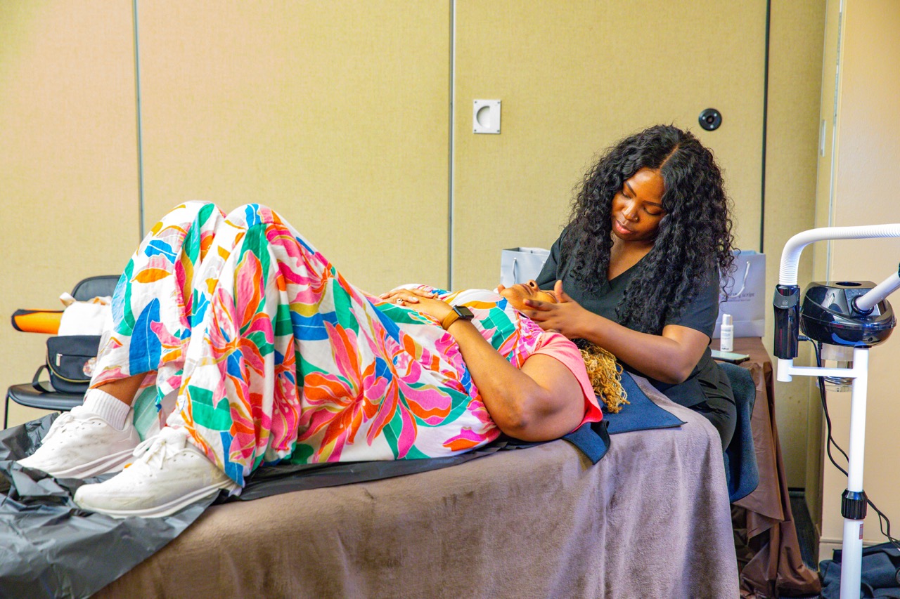 1W3A6288 Large - Community Impact Network Spa professional in dark scrubs treats a client's hands as the client lies on a padded table under a colorful blanket edge of a treatment room visible behind them, creating a manicure scene.