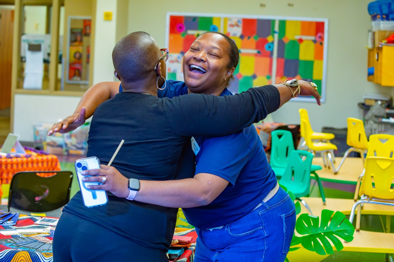 1W3A6375 Large - Community Impact Network Two women hug and laugh in a bright classroom; colorful chairs and a vibrant art display are in the background.