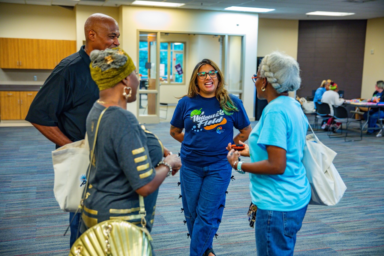 1W3A6570 Large - Community Impact Network Group of four adults in a circle talking and smiling in a bright community center hallway; one woman wears a blue Wellness Field shirt.