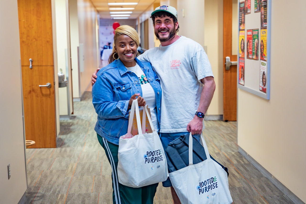 1W3A6588 Large - Community Impact Network Two smiling people stand in a hallway, each holding white tote bags that read'Rooted in Purpose' as they pose for a photo.