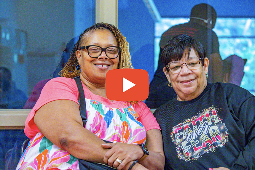 Screenshot - Community Impact Network Video thumbnail of two smiling women posing together; a red play button is centered over the image.