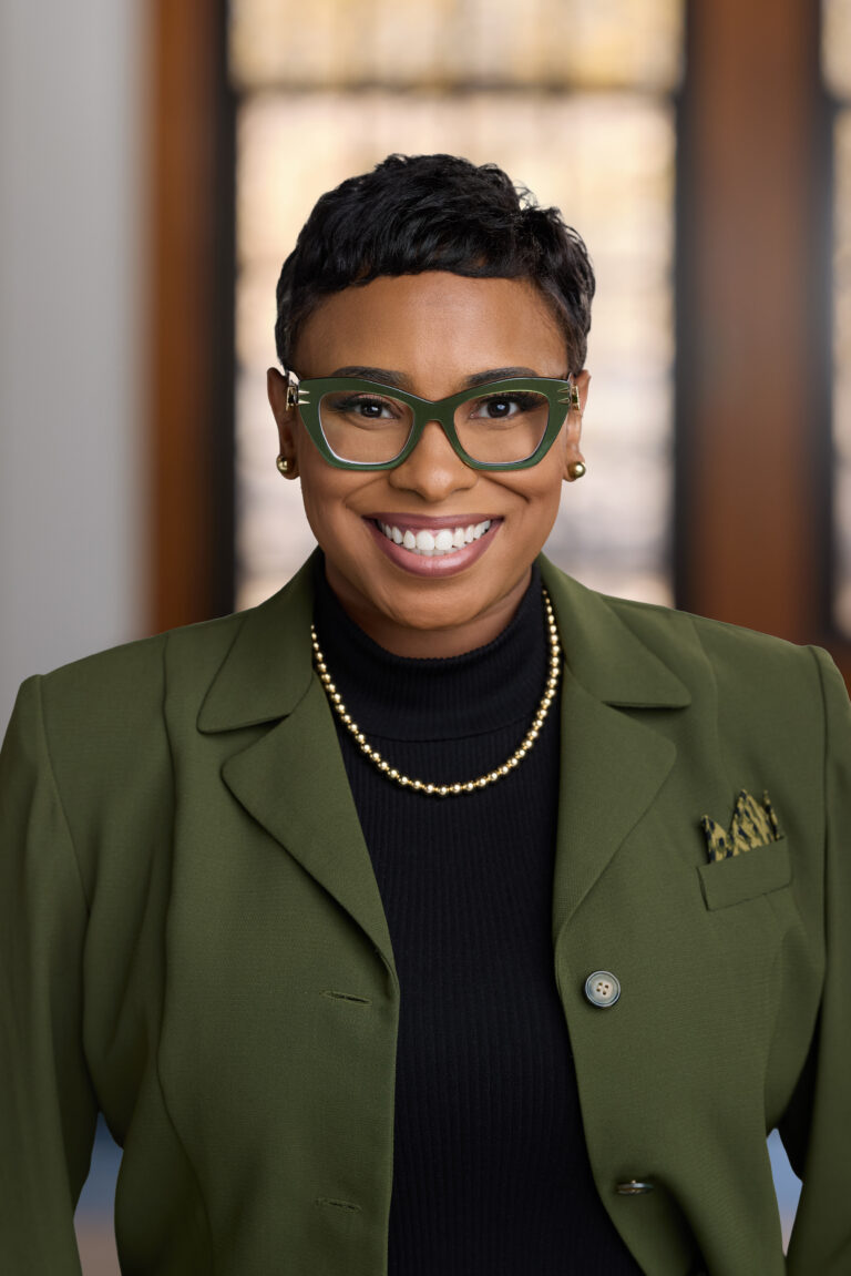 Smiling woman wearing a green blazer, black top, pearl necklace, and green cat-eye glasses.