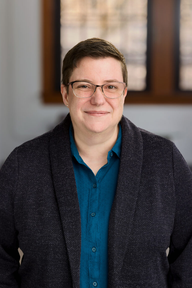 Smiling person with short dark hair and glasses, wearing a teal shirt and dark blazer in a room with windows behind.