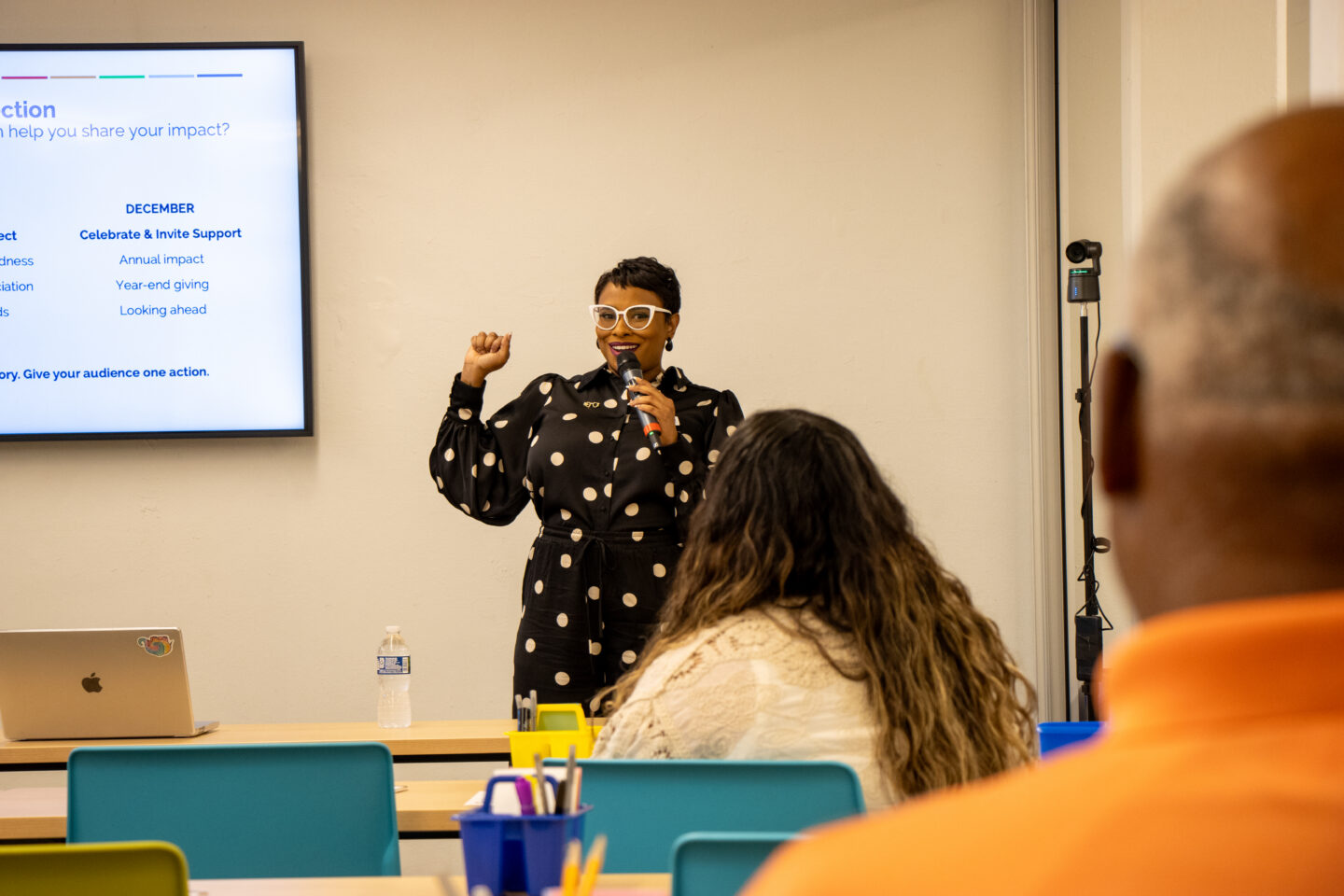 Female presenter in a black polka-dot dress and white glasses speaks into a microphone at the front of a classroom; a slide screen is on the left.